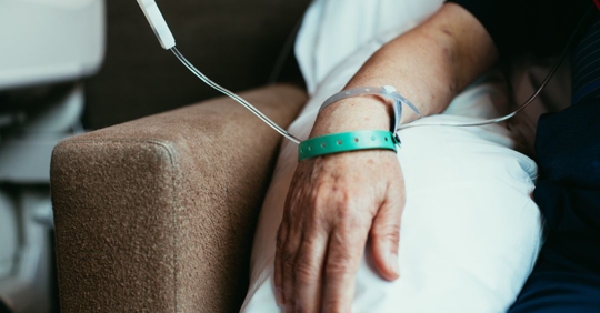 Close up photo of a person's arm resting on a pillow in a chair, with a green ID band, and a medical line.